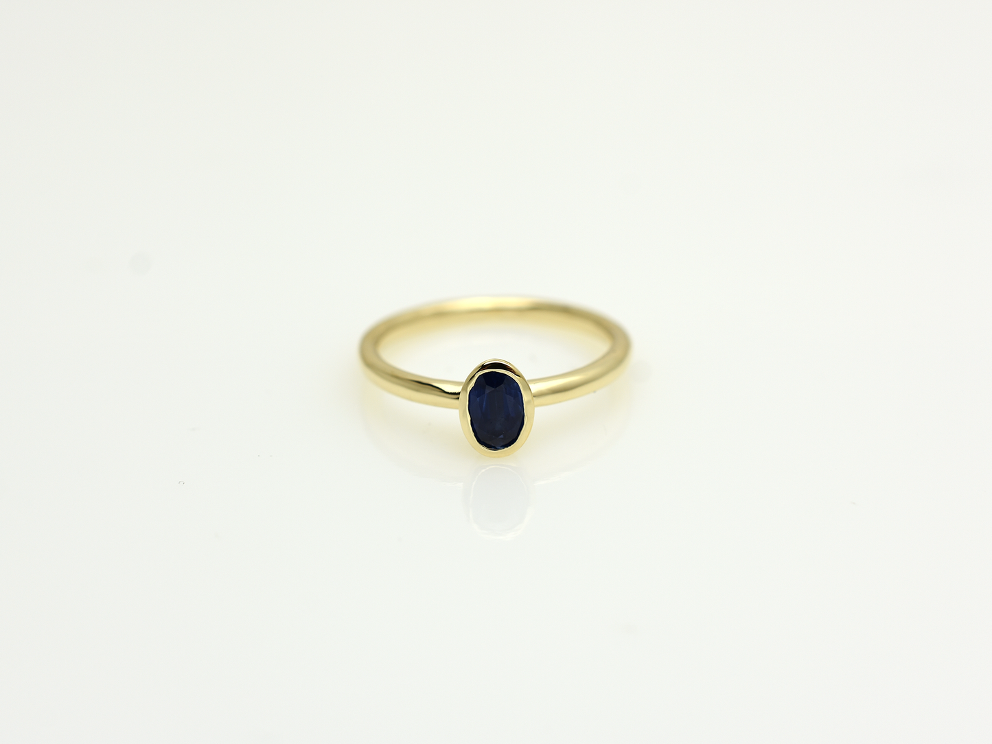 A minimalist 14k yellow gold solitaire ring featuring a 0.60 carat oval-cut deep blue sapphire. The gemstone is secured in a smooth bezel setting on a slim, polished gold band, centered against a clean white background with a soft reflection.