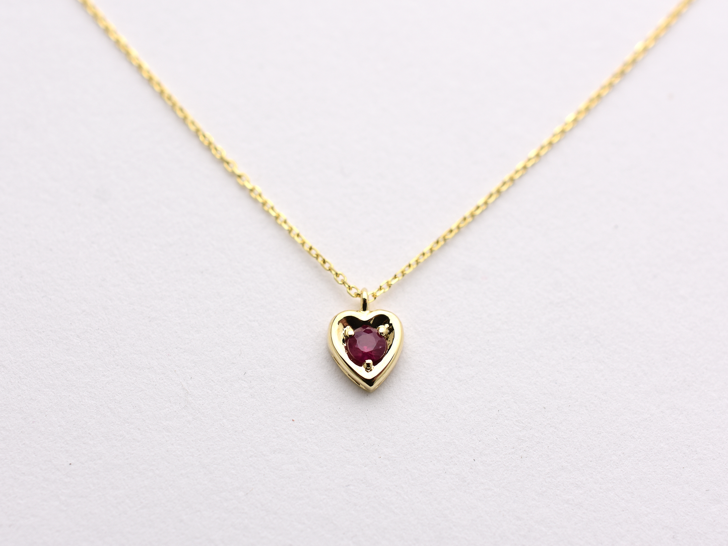 14k gold heart shaped necklace minimalistic design . in the center is a 3 mm natural red ruby , the chain is a 0.8mm dainty cable link chain displayed on a white background