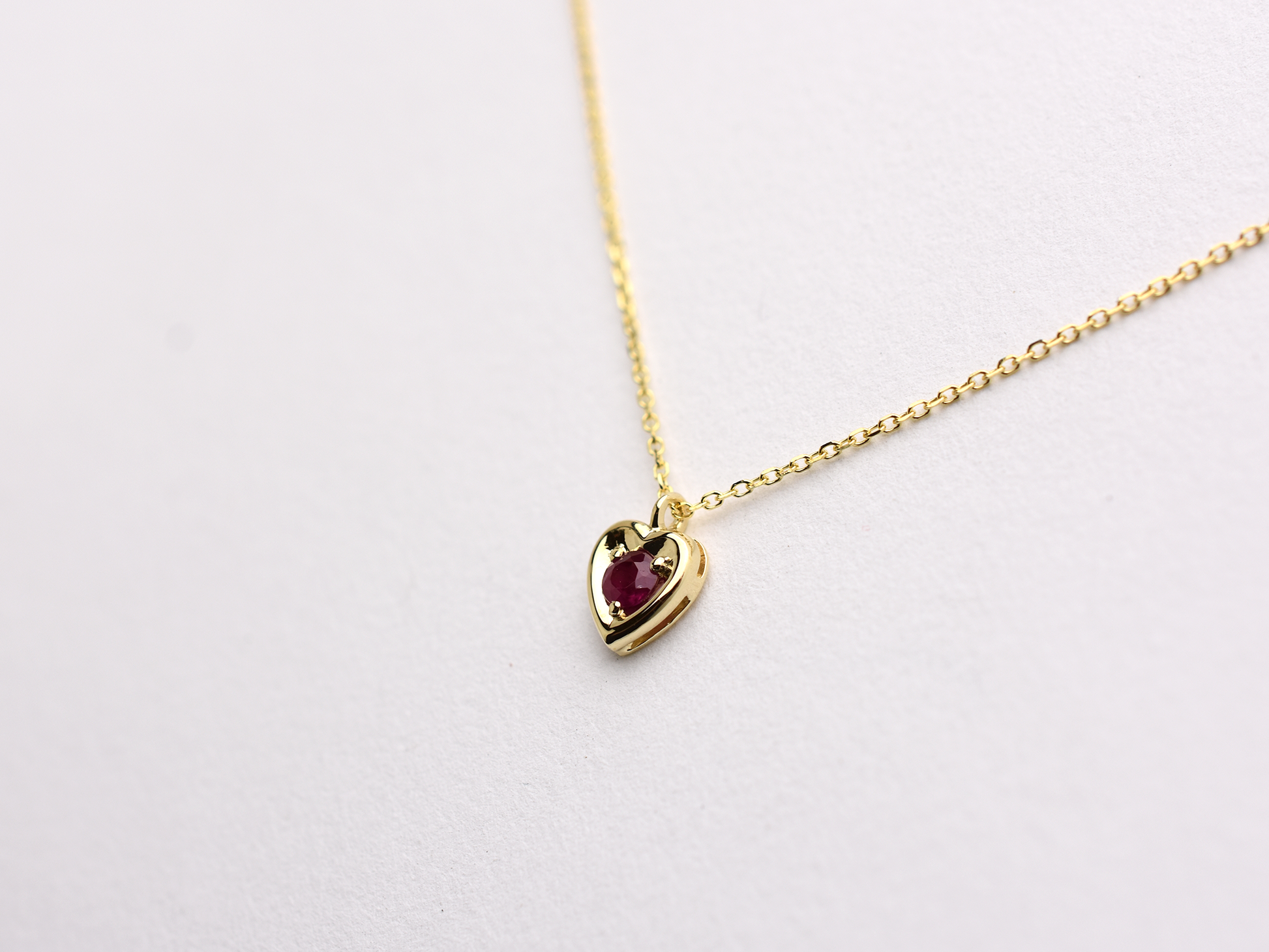 14k gold heart shaped necklace minimalistic design . in the center is a 3 mm natural red ruby , the chain is a 0.8mm dainty cable link chain displayed on a white background