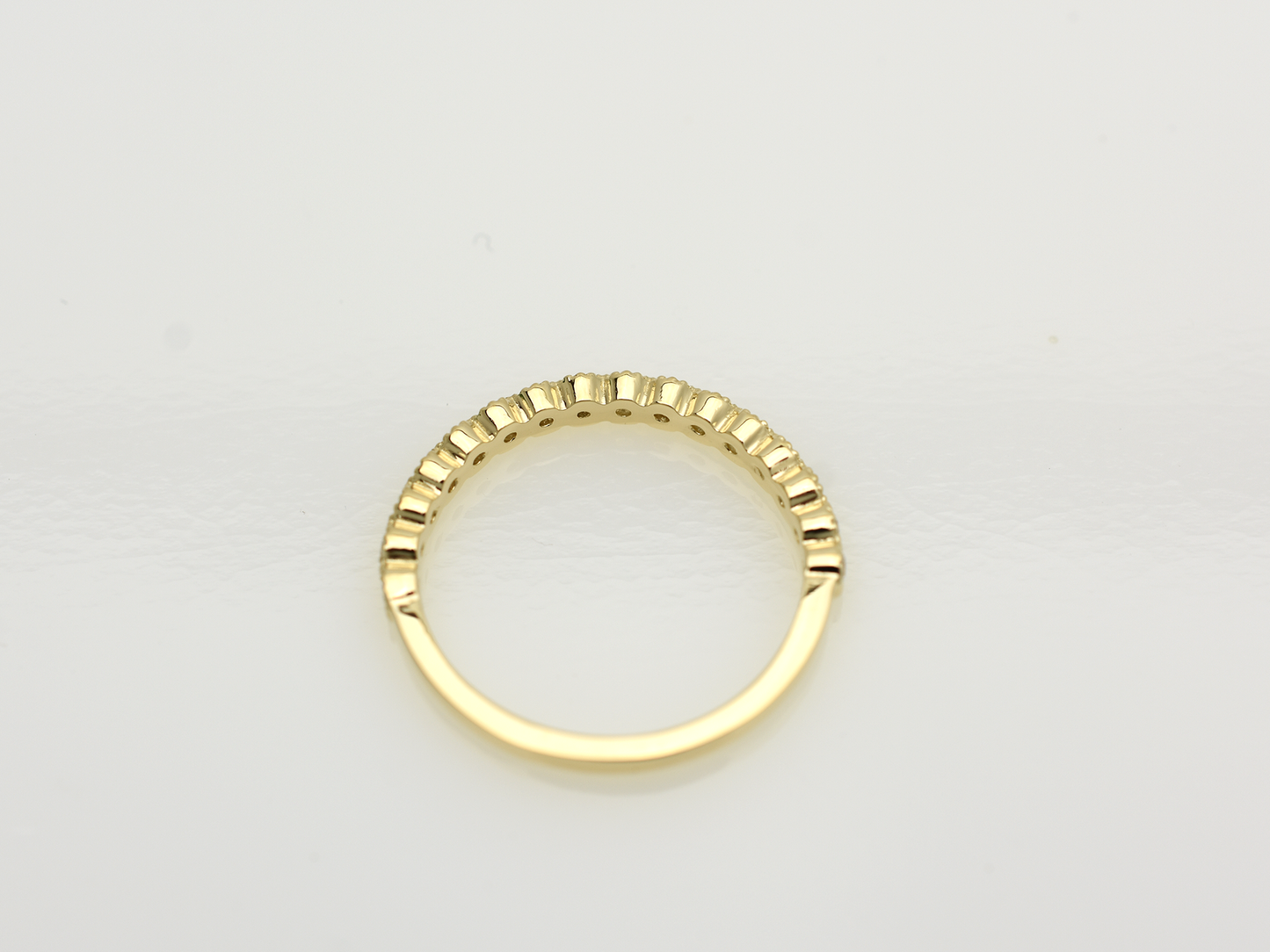 A close-up of a 14k yellow gold dainty wedding band featuring a row of round diamonds. Each diamond is set in a scalloped bezel with fine milgrain beaded edges. The ring is shown on a reflective white surface, highlighting the warm gold tones and sparkling stones.