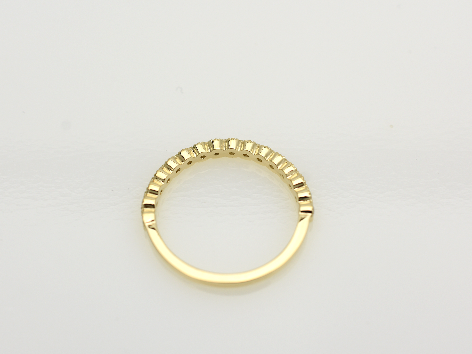 A close-up of a 14k yellow gold dainty wedding band featuring a row of round diamonds. Each diamond is set in a scalloped bezel with fine milgrain beaded edges. The ring is shown on a reflective white surface, highlighting the warm gold tones and sparkling stones.