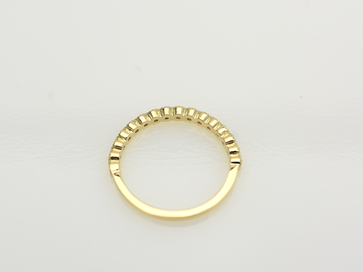 A close-up of a 14k yellow gold dainty wedding band featuring a row of round diamonds. Each diamond is set in a scalloped bezel with fine milgrain beaded edges. The ring is shown on a reflective white surface, highlighting the warm gold tones and sparkling stones.