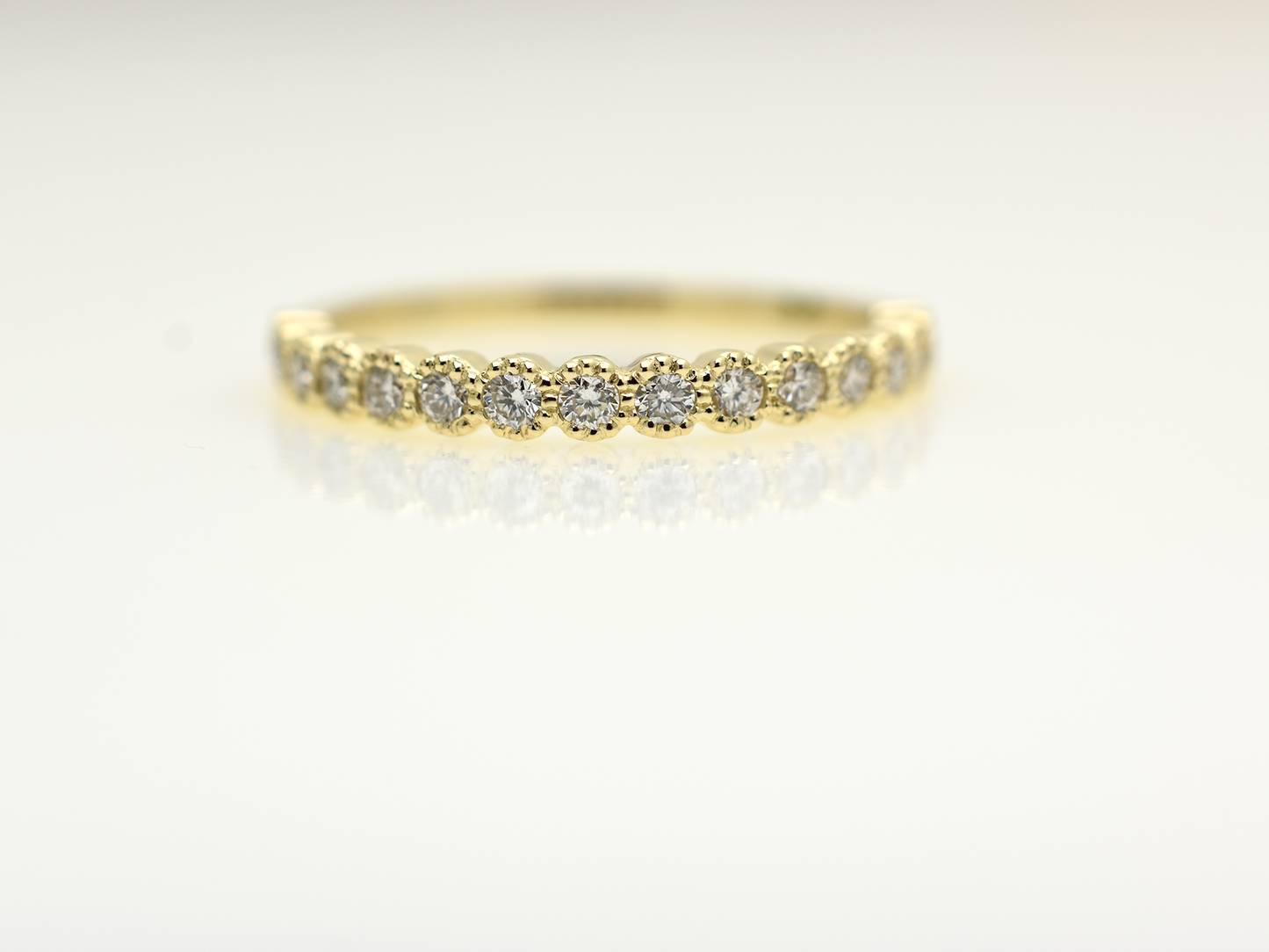 A close-up of a 14k yellow gold dainty wedding band featuring a row of round diamonds. Each diamond is set in a scalloped bezel with fine milgrain beaded edges. The ring is shown on a reflective white surface, highlighting the warm gold tones and sparkling stones.