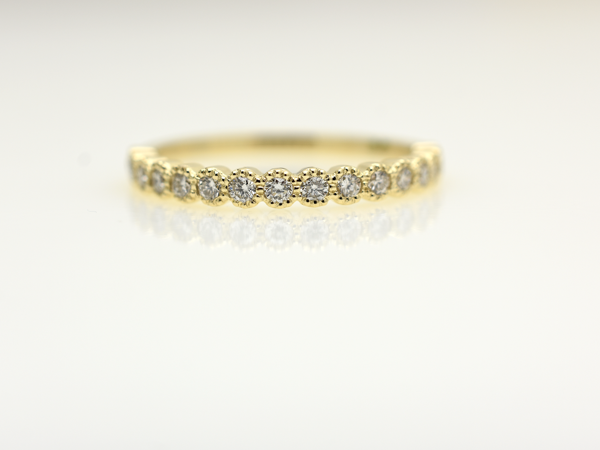 A close-up of a 14k yellow gold dainty wedding band featuring a row of round diamonds. Each diamond is set in a scalloped bezel with fine milgrain beaded edges. The ring is shown on a reflective white surface, highlighting the warm gold tones and sparkling stones.