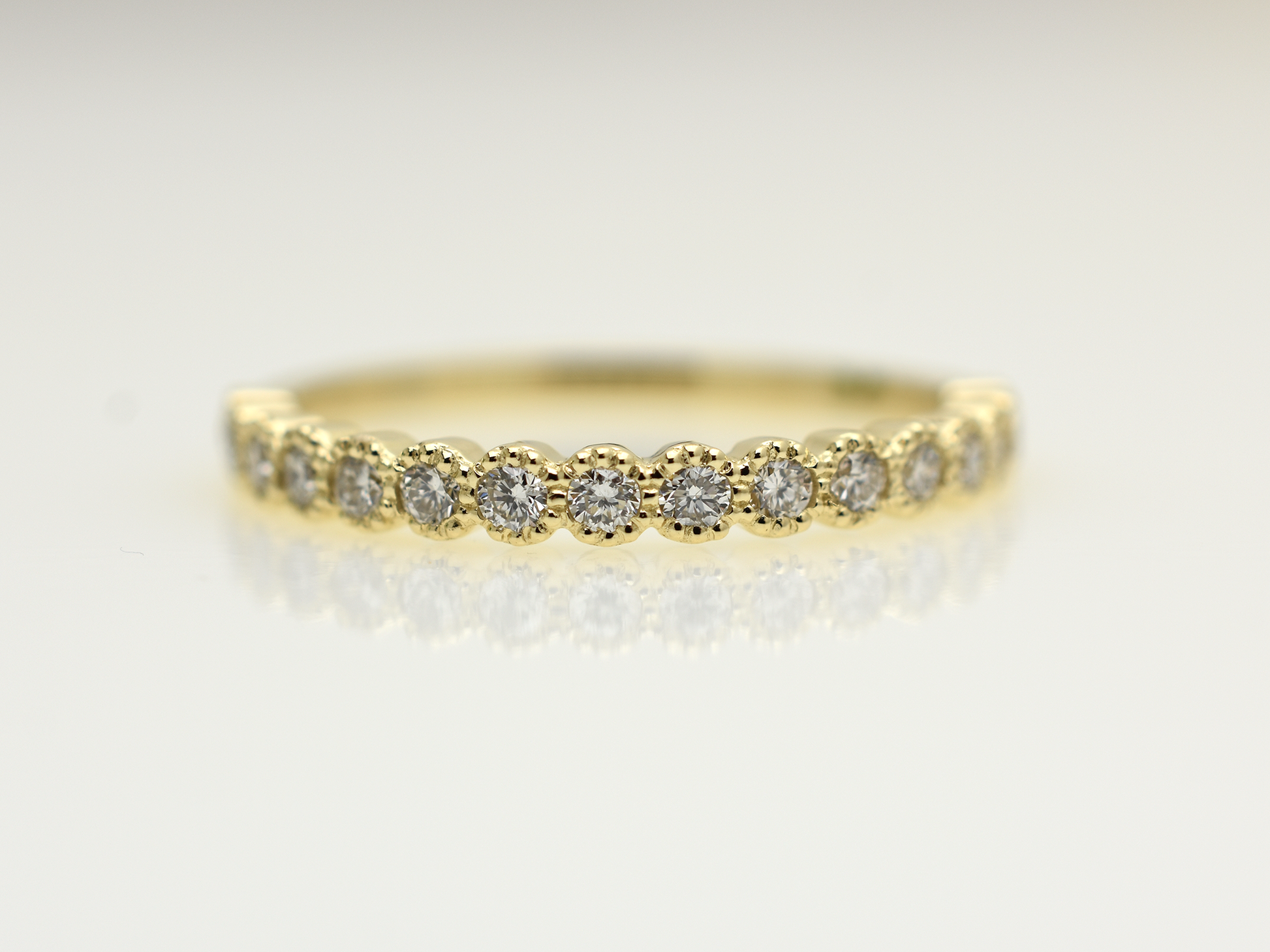 A close-up of a 14k yellow gold dainty wedding band featuring a row of round diamonds. Each diamond is set in a scalloped bezel with fine milgrain beaded edges. The ring is shown on a reflective white surface, highlighting the warm gold tones and sparkling stones.