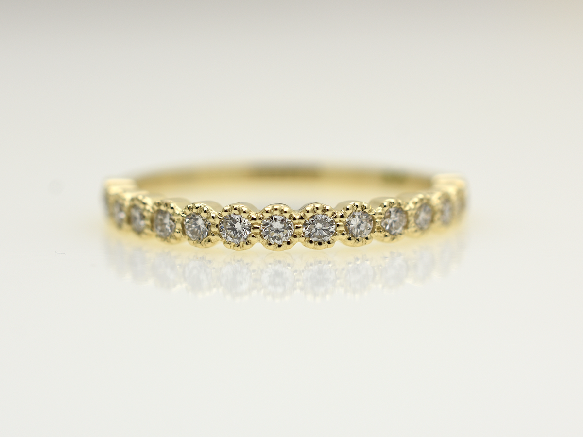 A close-up of a 14k yellow gold dainty wedding band featuring a row of round diamonds. Each diamond is set in a scalloped bezel with fine milgrain beaded edges. The ring is shown on a reflective white surface, highlighting the warm gold tones and sparkling stones.