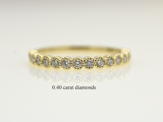 A close-up of a 14k yellow gold dainty wedding band featuring a row of round diamonds. Each diamond is set in a scalloped bezel with fine milgrain beaded edges. The ring is shown on a reflective white surface, highlighting the warm gold tones and sparkling stones.