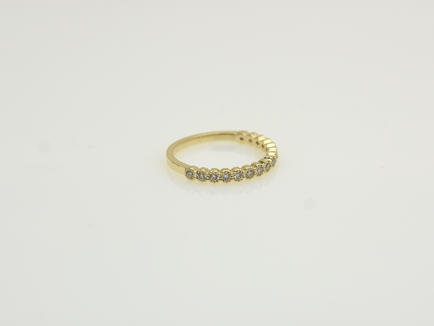 A close-up of a 14k yellow gold dainty wedding band featuring a row of round diamonds. Each diamond is set in a scalloped bezel with fine milgrain beaded edges. The ring is shown on a reflective white surface, highlighting the warm gold tones and sparkling stones.