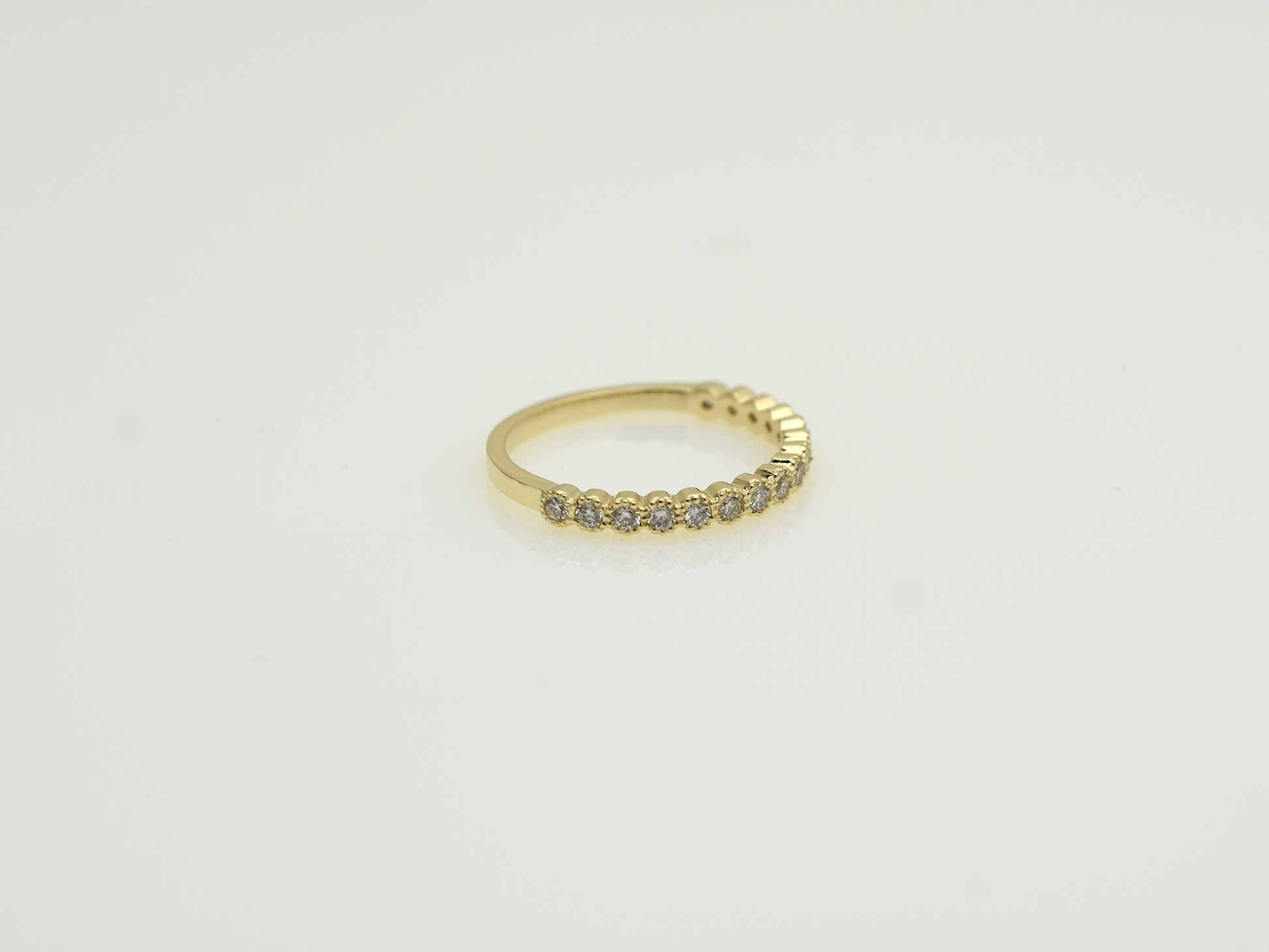 A close-up of a 14k yellow gold dainty wedding band featuring a row of round diamonds. Each diamond is set in a scalloped bezel with fine milgrain beaded edges. The ring is shown on a reflective white surface, highlighting the warm gold tones and sparkling stones.