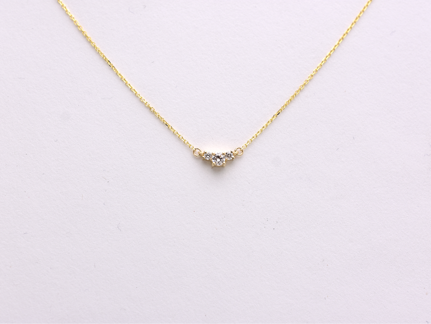  featuring a dainty three-stone diamond cluster pendant. The centerpiece consists of one slightly larger round brilliant cut stone flanked by two smaller stones in a minimalist prong setting on a fine gold cable chain with a lobster claw clasp