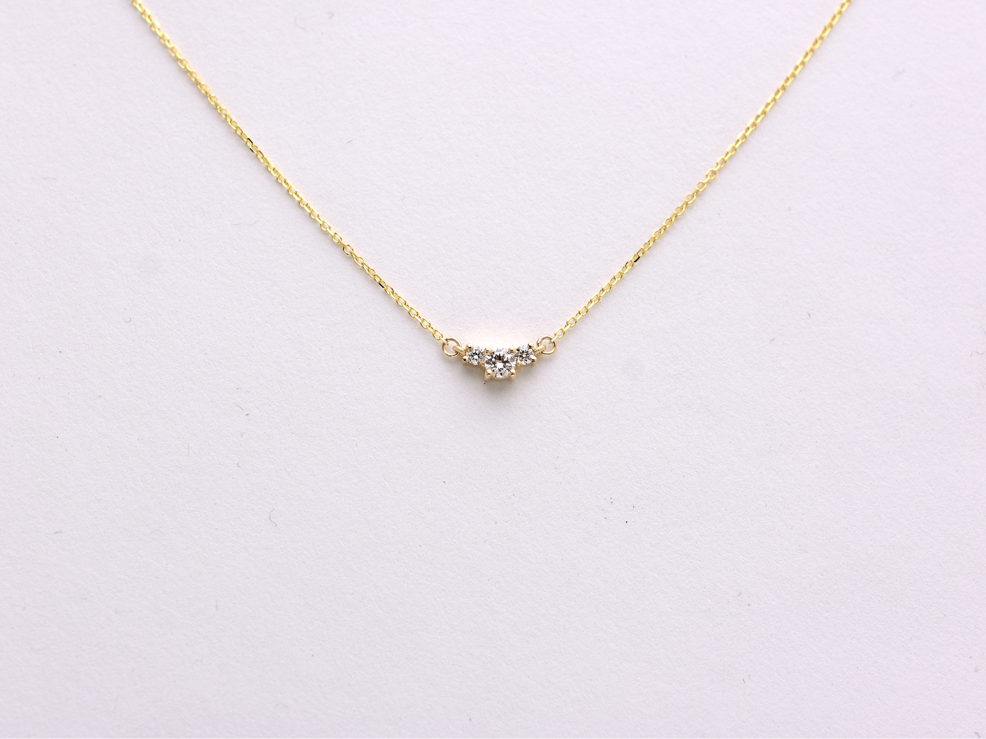  featuring a dainty three-stone diamond cluster pendant. The centerpiece consists of one slightly larger round brilliant cut stone flanked by two smaller stones in a minimalist prong setting on a fine gold cable chain with a lobster claw clasp