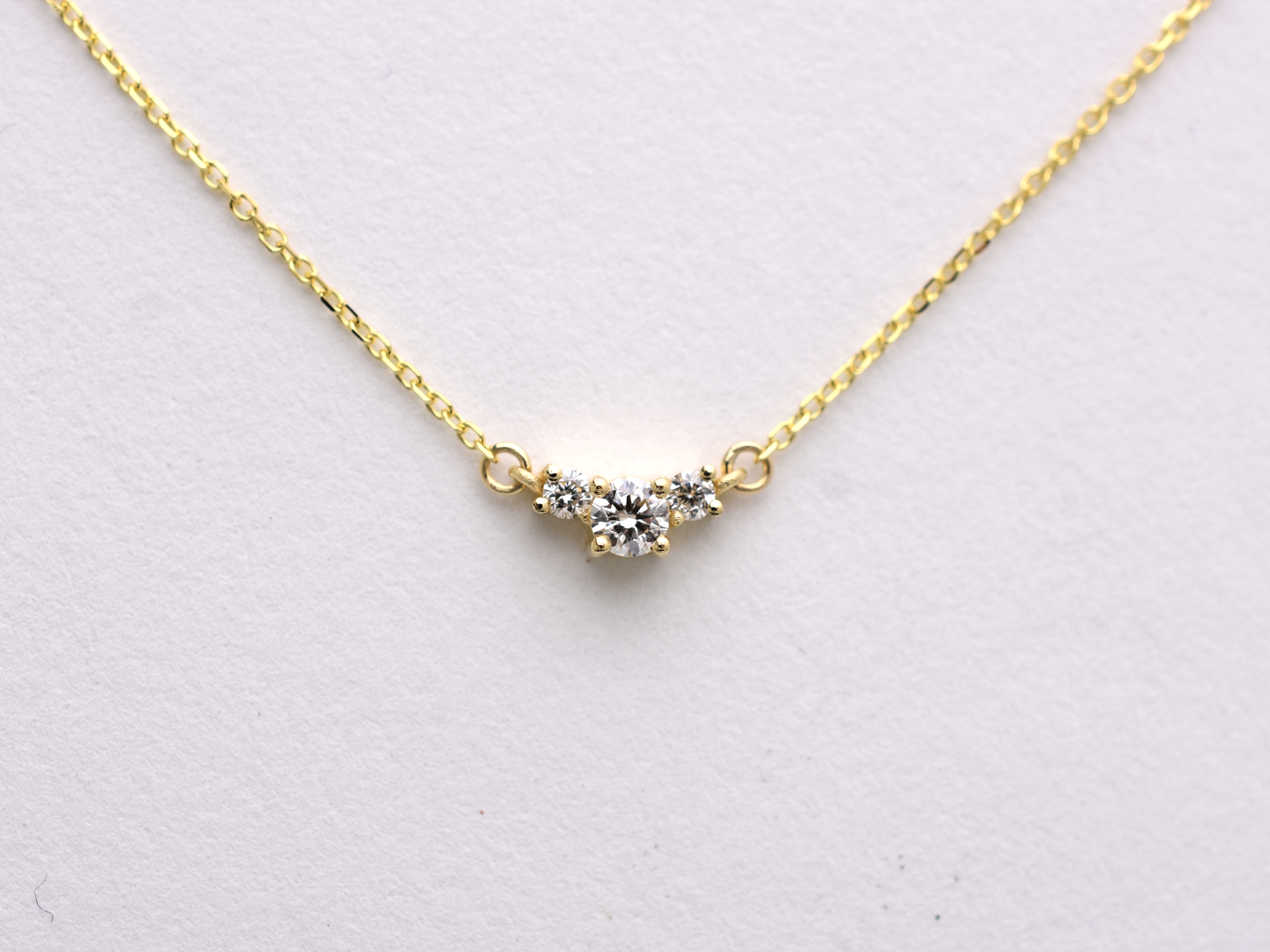 close up  featuring a dainty three-stone diamond cluster pendant. The centerpiece consists of one slightly larger round brilliant cut stone flanked by two smaller stones in a minimalist prong setting on a fine gold cable chain with a lobster claw clasp