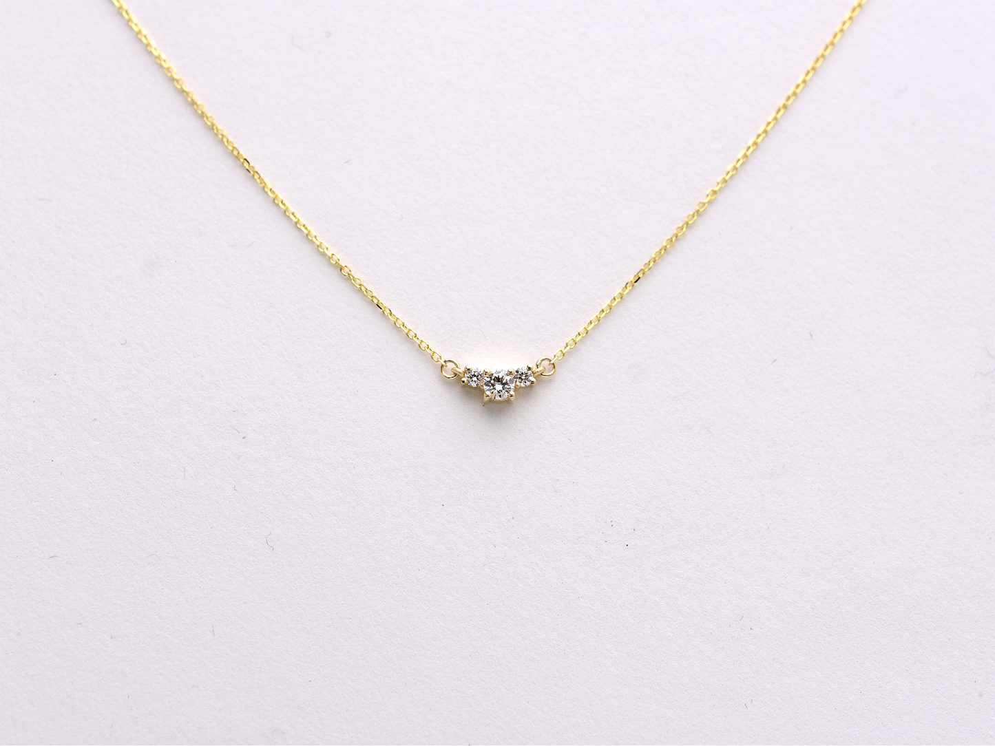  featuring a dainty three-stone diamond cluster pendant. The centerpiece consists of one slightly larger round brilliant cut stone flanked by two smaller stones in a minimalist prong setting on a fine gold cable chain with a lobster claw clasp