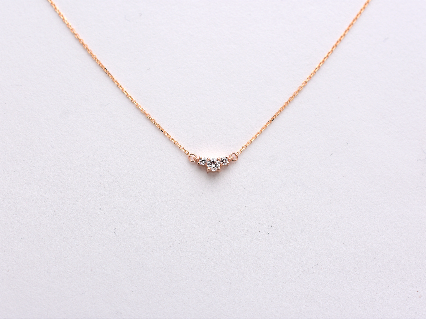  featuring a dainty three-stone diamond cluster pendant. The centerpiece consists of one slightly larger round brilliant cut stone flanked by two smaller stones in a minimalist prong setting on a fine gold cable chain with a lobster claw clasp
