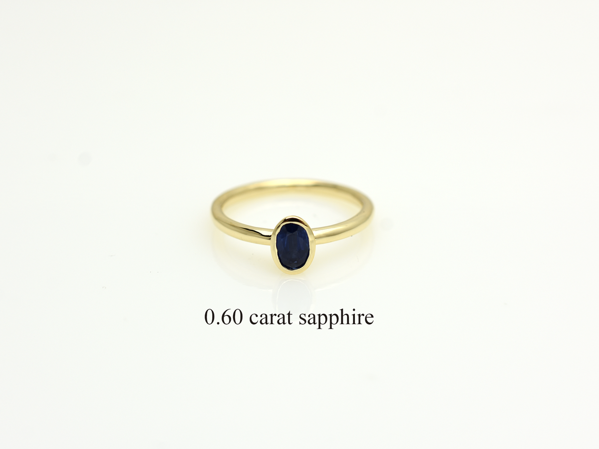 A minimalist 14k yellow gold solitaire ring featuring a 0.60 carat oval-cut deep blue sapphire. The gemstone is secured in a smooth bezel setting on a slim, polished gold band, centered against a clean white background with a soft reflection.