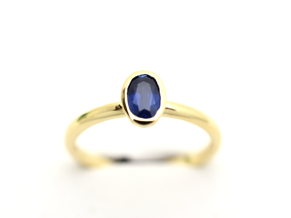 A minimalist 14k yellow gold solitaire ring featuring a 0.60 carat oval-cut deep blue sapphire. The gemstone is secured in a smooth bezel setting on a slim, polished gold band, centered against a clean white background with a soft reflection.