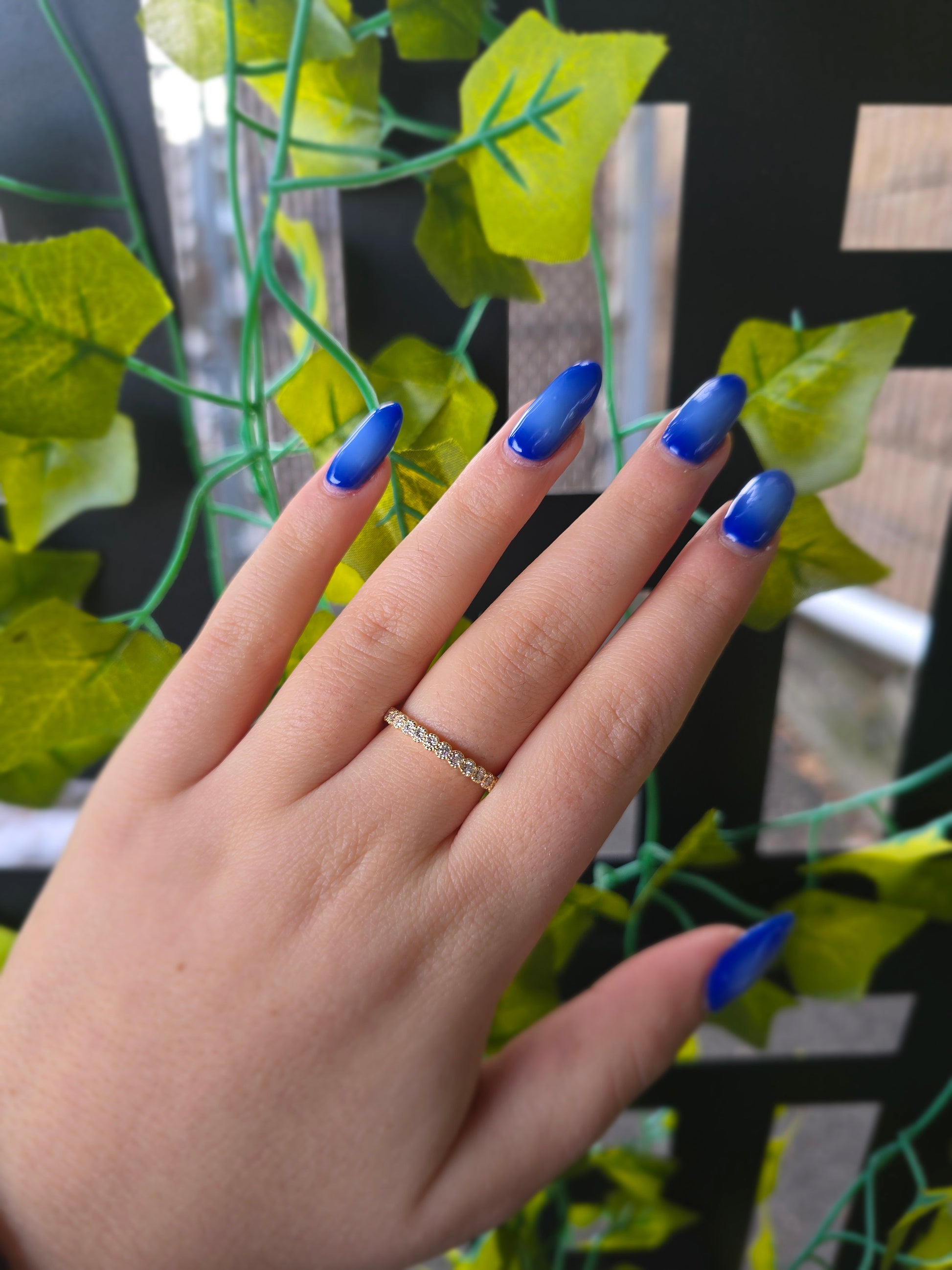 A close-up of a 14k yellow gold dainty wedding band featuring a row of round diamonds. Each diamond is set in a scalloped bezel with fine milgrain beaded edges. The ring is shown on a reflective white surface, highlighting the warm gold tones and sparkling stones.
