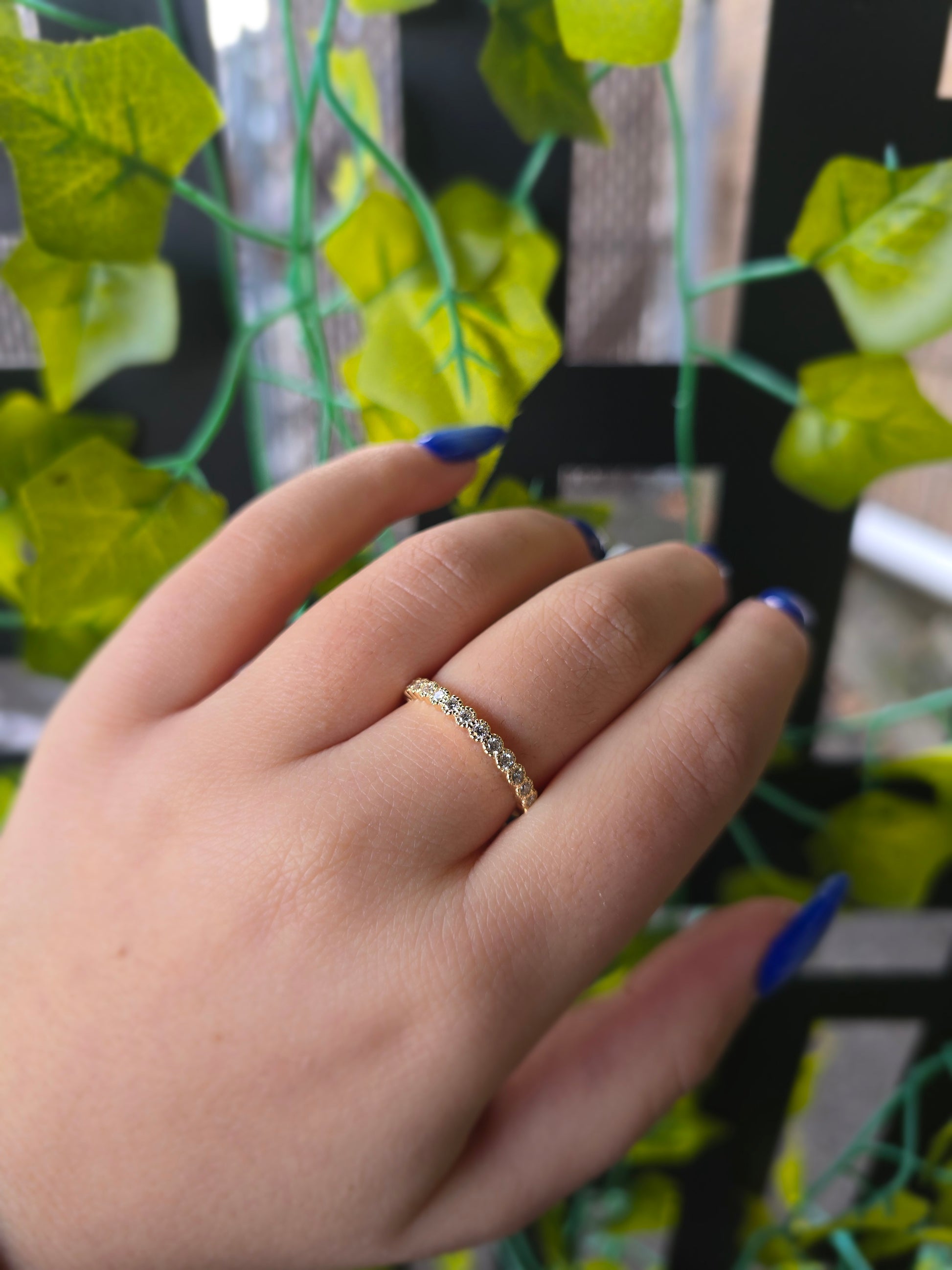 A close-up of a 14k yellow gold dainty wedding band featuring a row of round diamonds. Each diamond is set in a scalloped bezel with fine milgrain beaded edges. The ring is shown on a reflective white surface, highlighting the warm gold tones and sparkling stones.