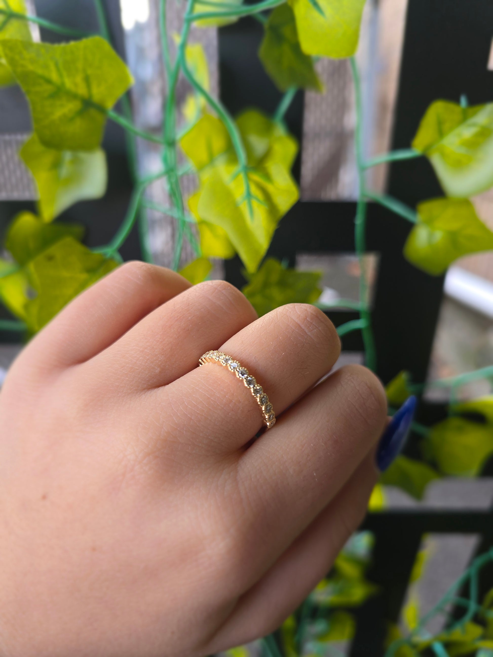 A close-up of a 14k yellow gold dainty wedding band featuring a row of round diamonds. Each diamond is set in a scalloped bezel with fine milgrain beaded edges. The ring is shown on a reflective white surface, highlighting the warm gold tones and sparkling stones.