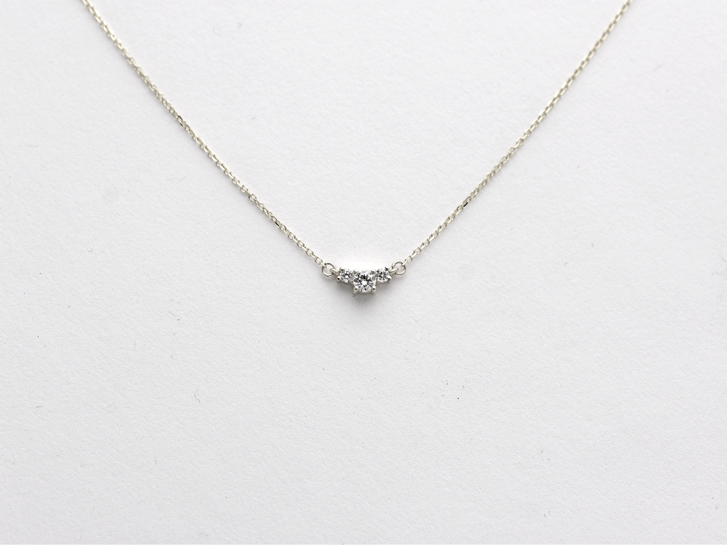  featuring a dainty three-stone diamond cluster pendant. The centerpiece consists of one slightly larger round brilliant cut stone flanked by two smaller stones in a minimalist prong setting on a fine gold cable chain with a lobster claw clasp