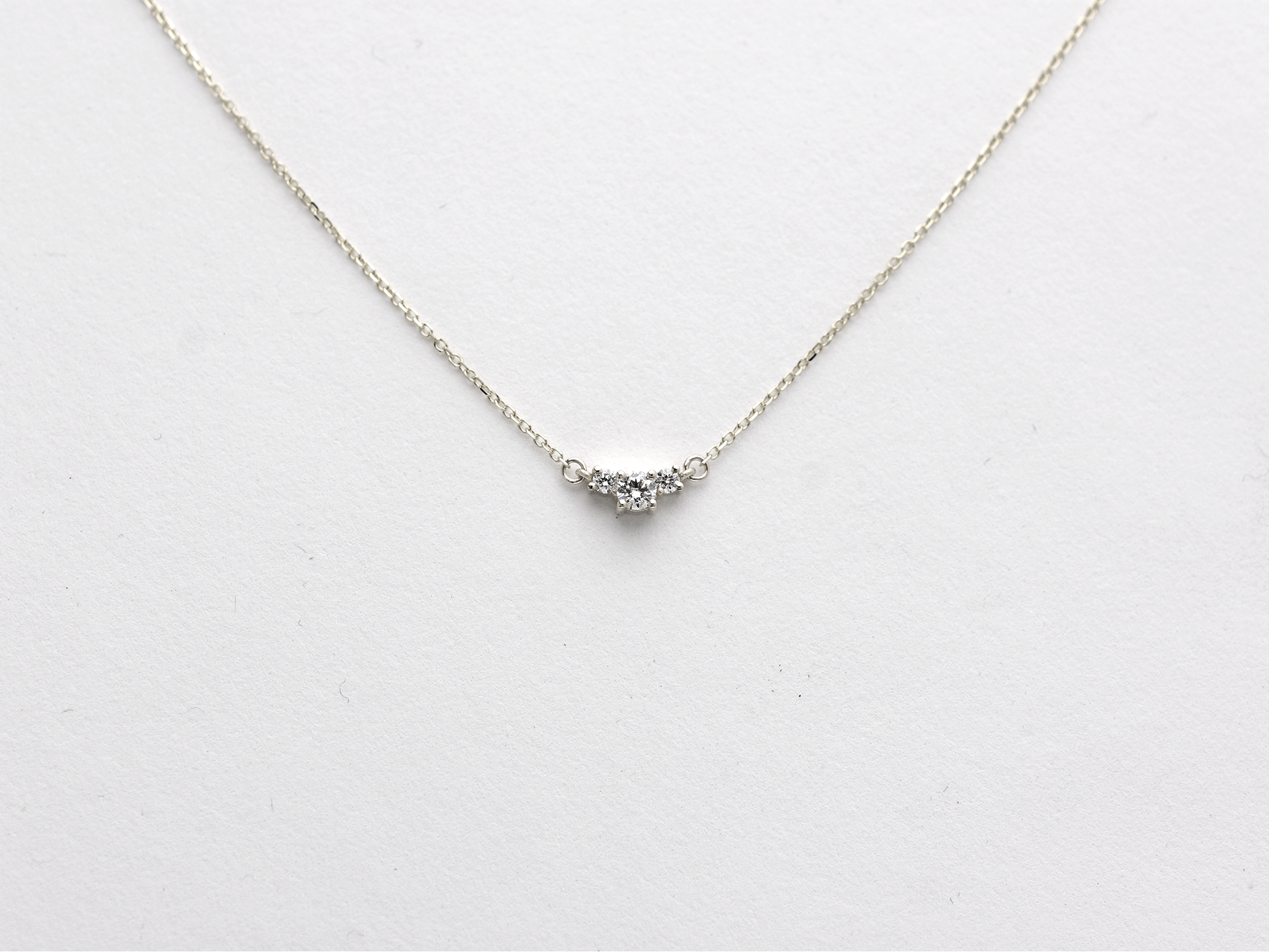  featuring a dainty three-stone diamond cluster pendant. The centerpiece consists of one slightly larger round brilliant cut stone flanked by two smaller stones in a minimalist prong setting on a fine gold cable chain with a lobster claw clasp