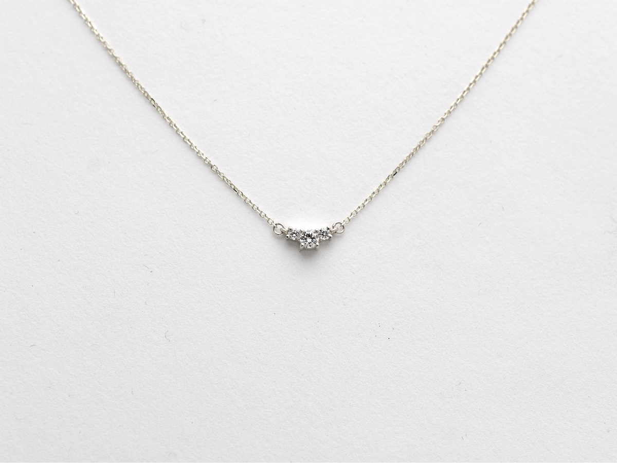  featuring a dainty three-stone diamond cluster pendant. The centerpiece consists of one slightly larger round brilliant cut stone flanked by two smaller stones in a minimalist prong setting on a fine gold cable chain with a lobster claw clasp