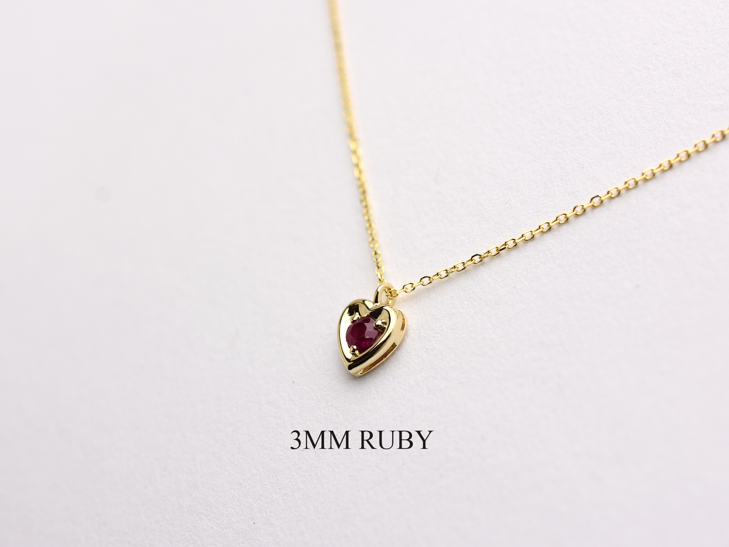 14k gold heart shaped necklace minimalistic design . in the center is a 3 mm natural red ruby , the chain is a 0.8mm dainty cable link chain displayed on a white background