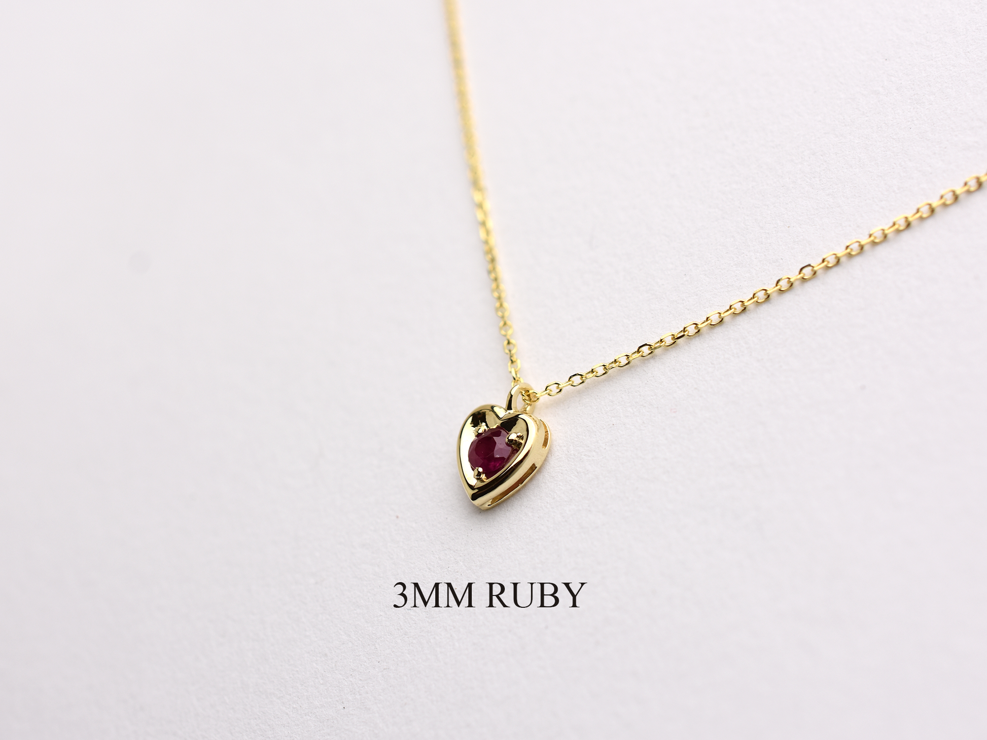 14k gold heart shaped necklace minimalistic design . in the center is a 3 mm natural red ruby , the chain is a 0.8mm dainty cable link chain displayed on a white background