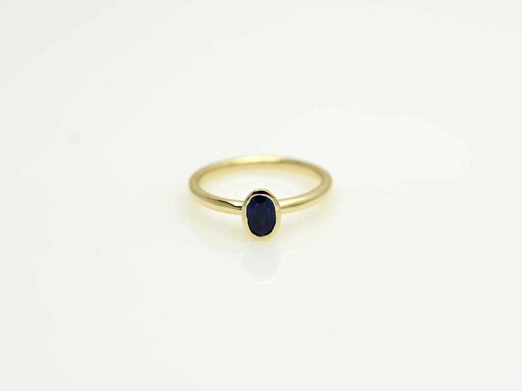 A minimalist 14k yellow gold solitaire ring featuring a 0.60 carat oval-cut deep blue sapphire. The gemstone is secured in a smooth bezel setting on a slim, polished gold band, centered against a clean white background with a soft reflection.
