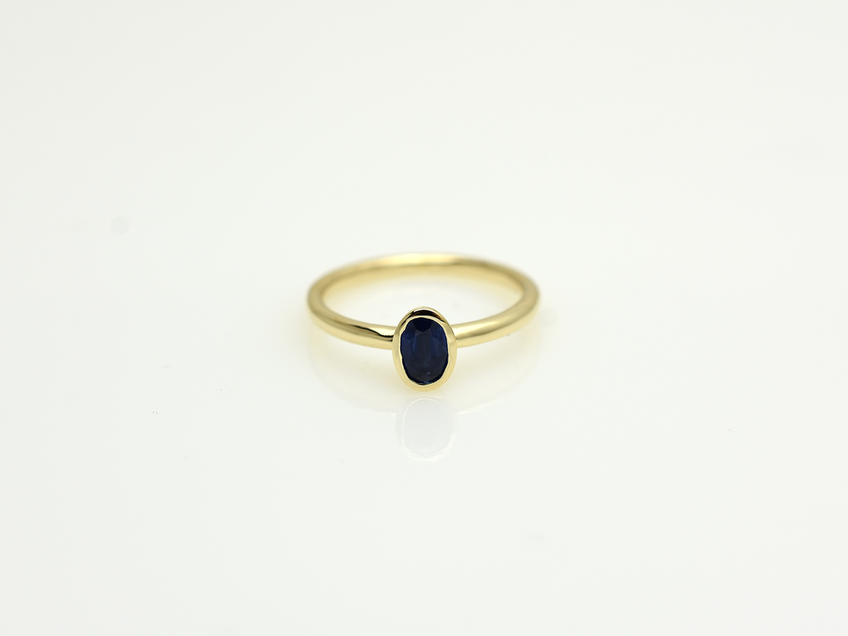 A minimalist 14k yellow gold solitaire ring featuring a 0.60 carat oval-cut deep blue sapphire. The gemstone is secured in a smooth bezel setting on a slim, polished gold band, centered against a clean white background with a soft reflection.