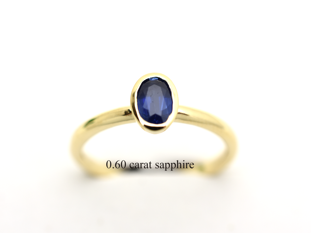 A minimalist 14k yellow gold solitaire ring featuring a 0.60 carat oval-cut deep blue sapphire. The gemstone is secured in a smooth bezel setting on a slim, polished gold band, centered against a clean white background with a soft reflection.