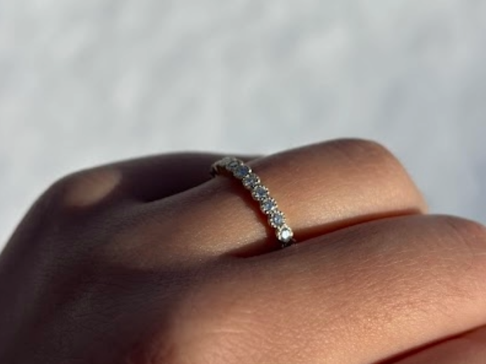 A close-up of a 14k yellow gold dainty wedding band featuring a row of round diamonds. Each diamond is set in a scalloped bezel with fine milgrain beaded edges. The ring is shown on a reflective white surface, highlighting the warm gold tones and sparkling stones.