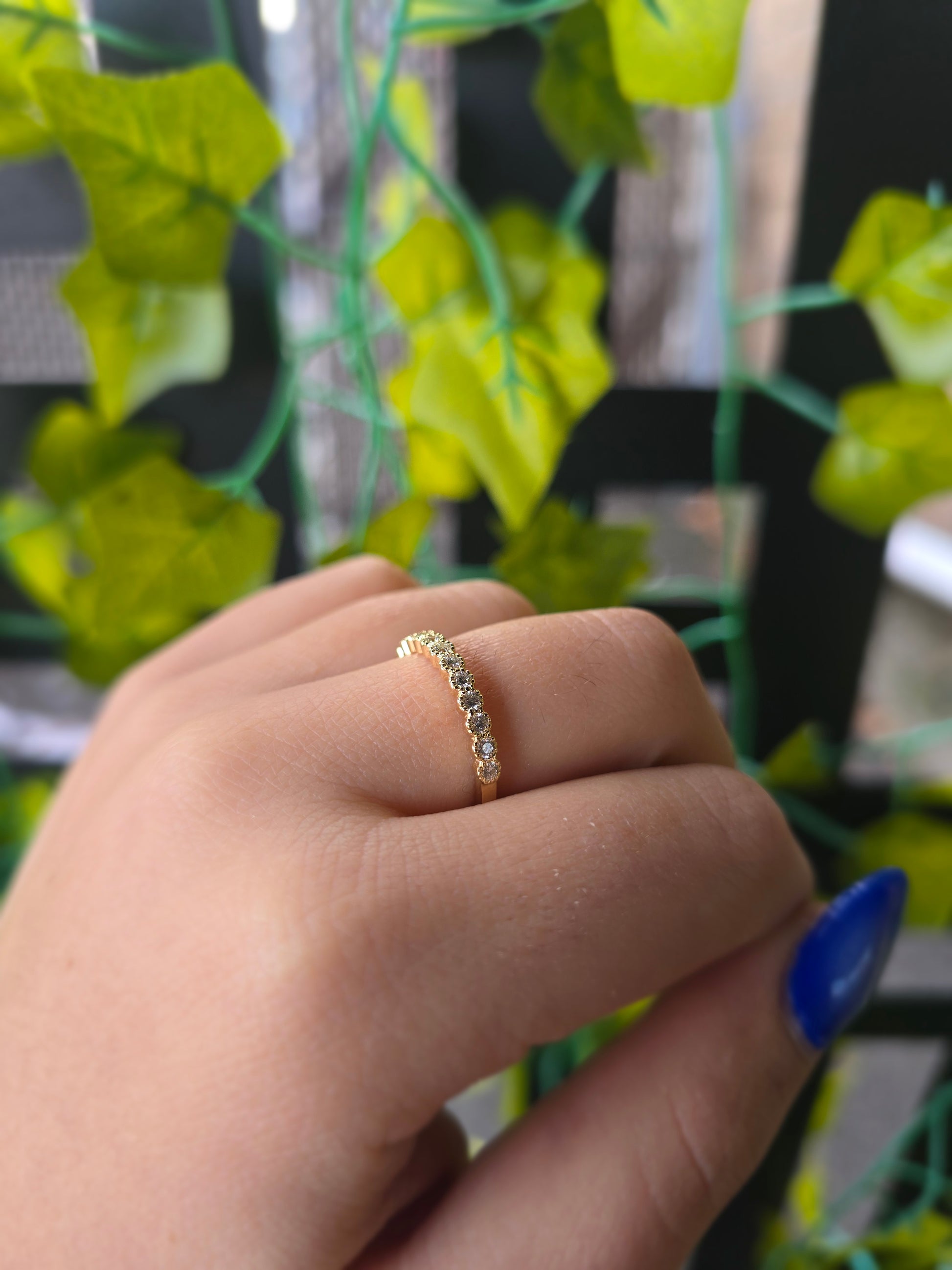 14k yellow gold dainty wedding band featuring a row of round diamonds. Each diamond is set in a scalloped bezel with fine milgrain beaded edges. The ring is shown on a reflective white surface, highlighting the warm gold tones and sparkling stones.