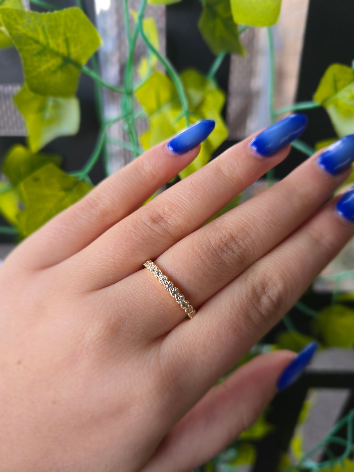  14k yellow gold dainty wedding band featuring a row of round diamonds. Each diamond is set in a scalloped bezel with fine milgrain beaded edges. The ring is shown on a reflective white surface, highlighting the warm gold tones and sparkling stones.