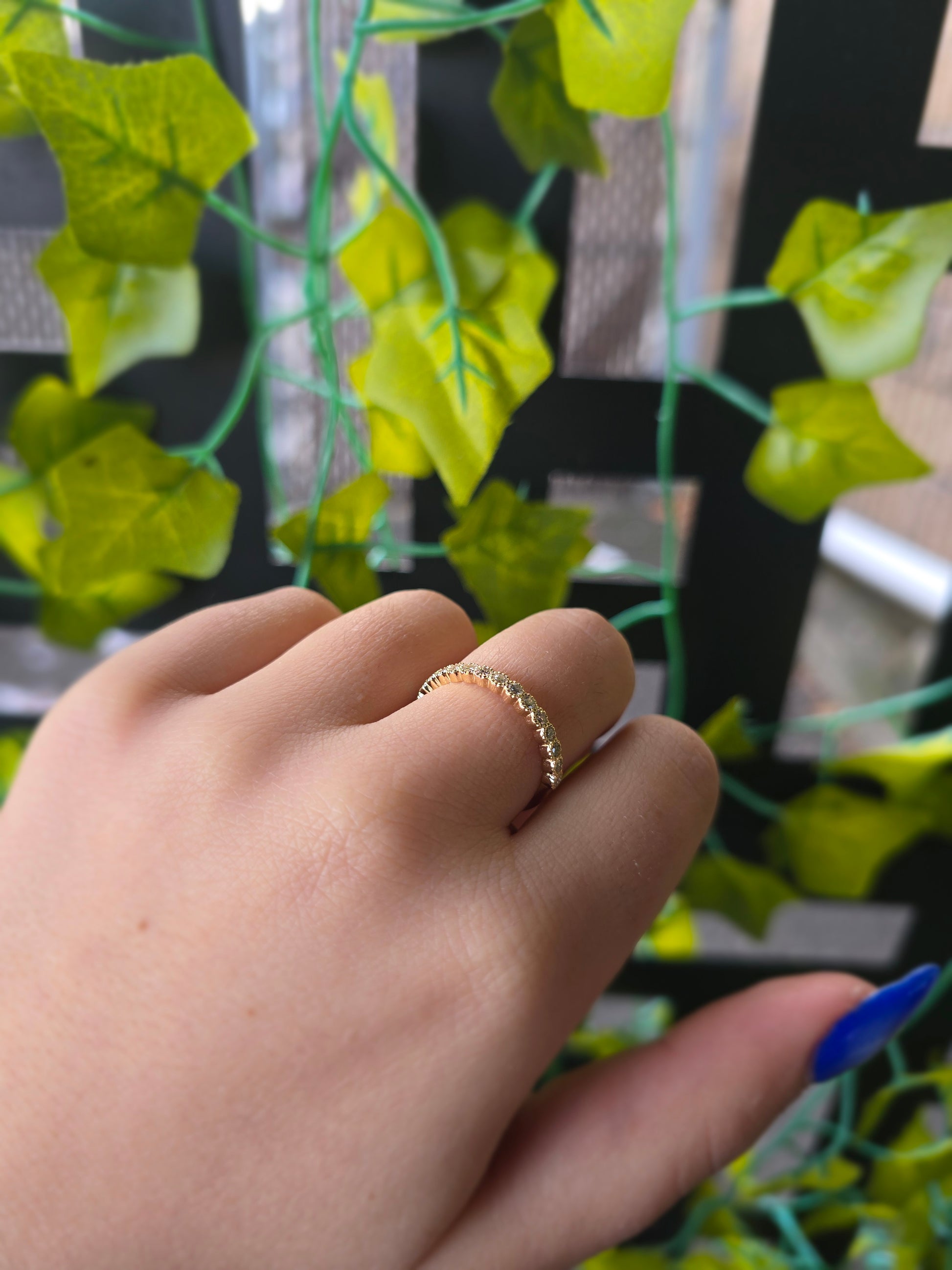 A close-up of a 14k yellow gold dainty wedding band featuring a row of round diamonds. Each diamond is set in a scalloped bezel with fine milgrain beaded edges. The ring is shown on a reflective white surface, highlighting the warm gold tones and sparkling stones.
